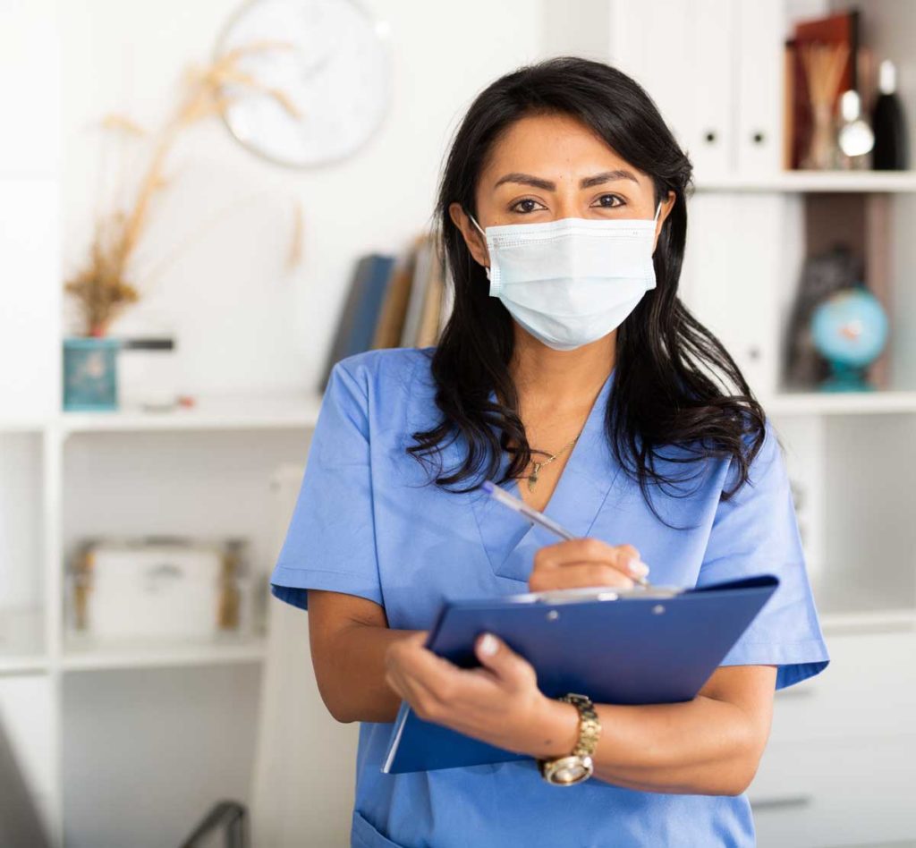 Portrait of latin american female doctor in surgical face mask meeting patient in medical office, filling out medical form at clipboard